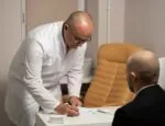 A bald physician writes notes on a piece of paper. Another man sits across from the physician looking at him.