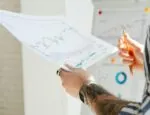 A person holds a piece of paper with various line graphs all trending upward. Only their arms are in the frame and there are more line graphs behind them.