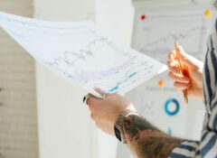 A person holds a piece of paper with various line graphs all trending upward. Only their arms are in the frame and there are more line graphs behind them.